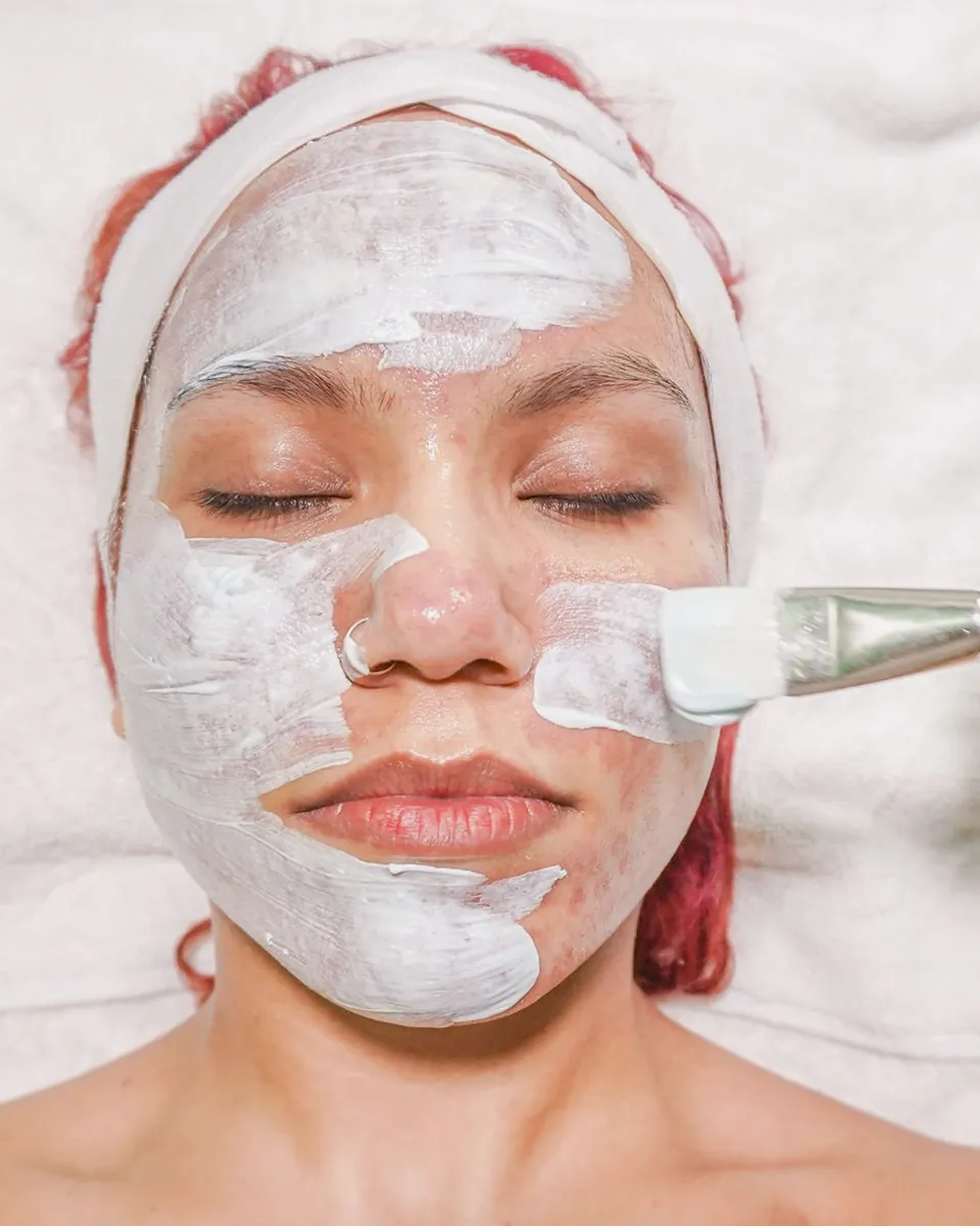 A white mask being brushed onto a woman's face during a facial.