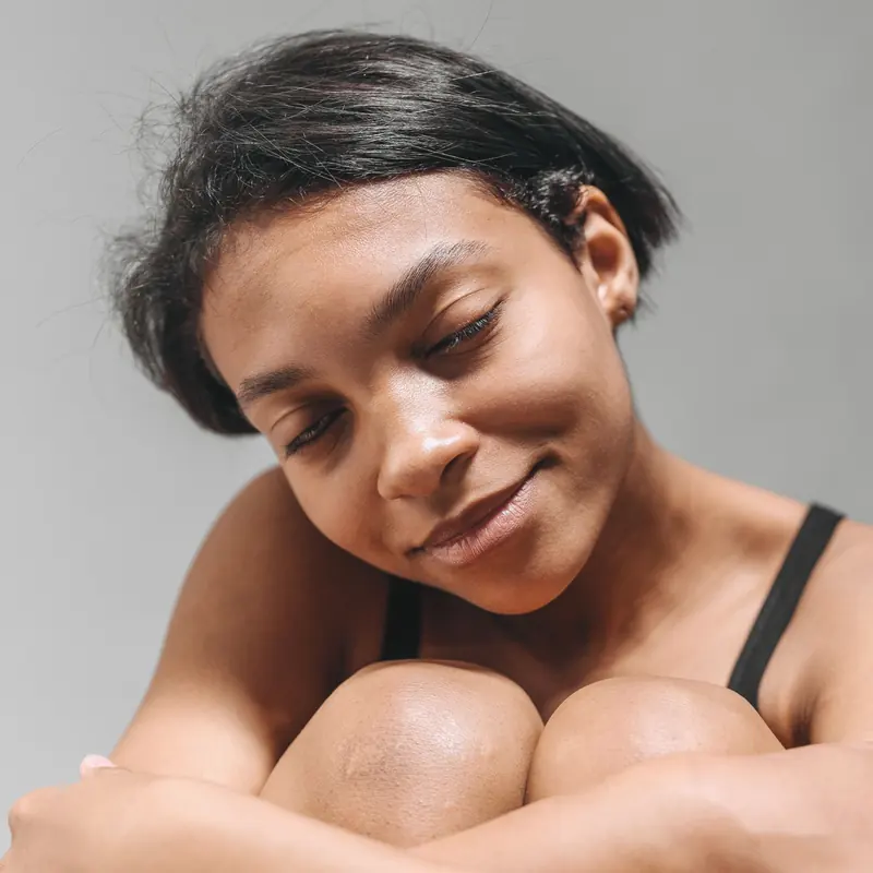 A woman with short dark hair resting her cheek on her knees, eyes closed and smiling