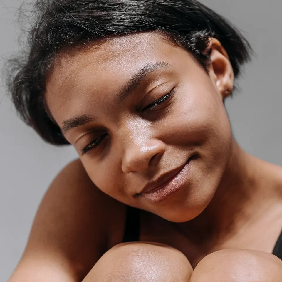 Close-up of a woman with her eyes closed and a slight smile, her head tilted over her knees, against a pale grey background.