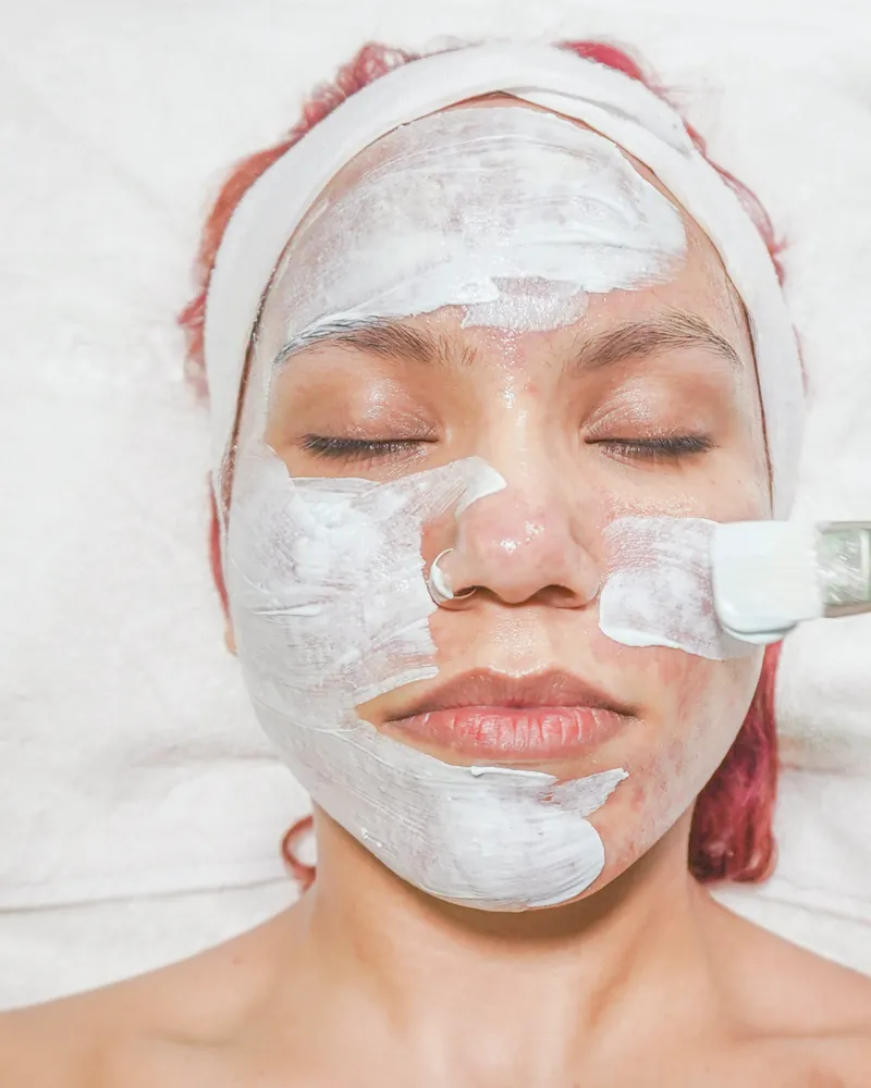A brush spreading a white mask over a woman’s face while her eyes are closed