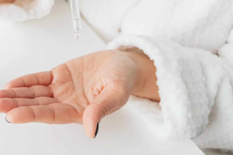 An open hand held out beneath a glass dropper, beside the sleeve of a white robe
